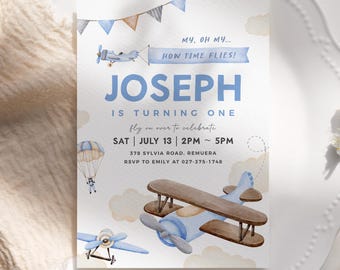 May include: A blue and brown airplane with a banner that says "How Time Flies!"  A blue and white parachute with a small figure in it.  The text "Joseph is turning one" is in blue and white.  The text "fly on over to celebrate" is in black.  The text "Sat July 13 2PM - 5PM" is in black.  The text "378 Sylvia Road, Remuera" is in black.  The text "RSVP to Emily at 027-375-1748" is in black.