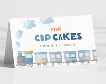 May include: A white card with the words "MINI CUP CAKES" in blue and orange. Below the text is "BLUEBERRY & CHOCOLATE". A cartoon train with blue and tan cars is at the bottom, with blue clouds in the background.
