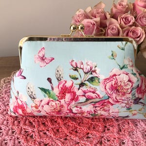 May include: A pale blue clutch bag with a gold clasp, featuring a floral design of pink roses and butterflies. The bag rests on a pink crocheted surface, with a bouquet of pink roses in the background.