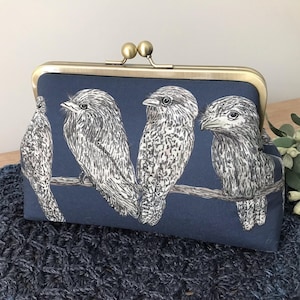 May include: A navy blue fabric clutch purse featuring a black and white line art illustration of four birds perched on a branch. The purse has a metallic brass-toned frame with a double ball clasp closure at the top for secure opening and closing.