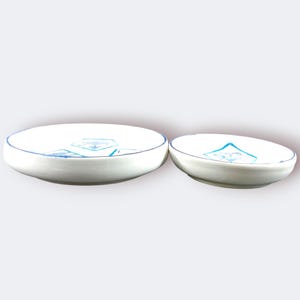 May include: Two white ceramic bowls with blue trim and a blue geometric design. The bowls are different sizes, with the larger one on the left and the smaller one on the right. The bowls are likely for serving or display.