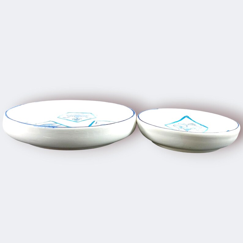 May include: Two white ceramic bowls with blue trim and a blue geometric design. The bowls are different sizes, with the larger one on the left and the smaller one on the right. The bowls are likely for serving or display.