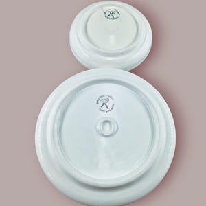 May include: Two white ceramic lids, one larger than the other, are displayed against a muted pink background. The underside of each lid features a central knob and a circular logo with text. The lids appear to be part of a pottery set.