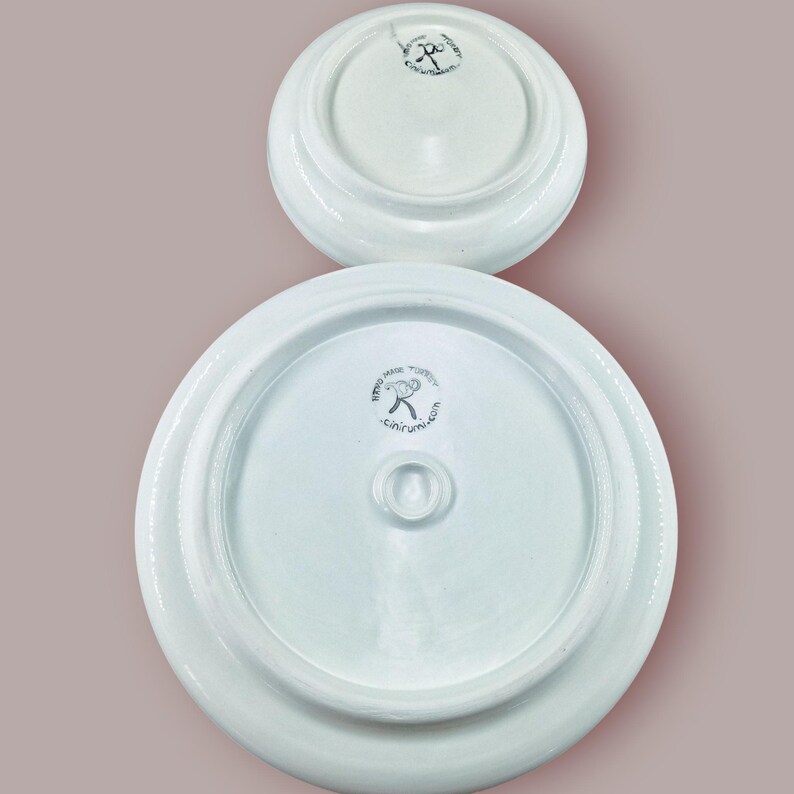 May include: Two white ceramic lids, one larger than the other, are displayed against a muted pink background. The underside of each lid features a central knob and a circular logo with text. The lids appear to be part of a pottery set.