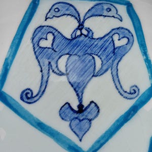 May include: Close-up of a white ceramic surface with a blue geometric design. The design features a central stylized motif with heart-shaped elements and bird-like figures, framed by a blue border.