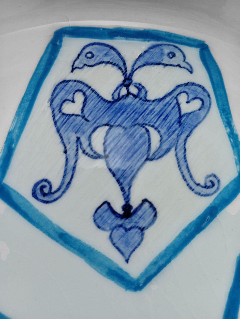 May include: Close-up of a white ceramic surface with a blue geometric design. The design features a central stylized motif with heart-shaped elements and bird-like figures, framed by a blue border.