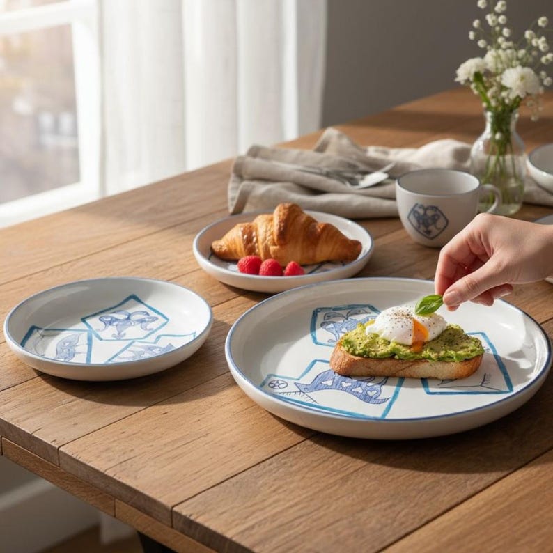 May include: White ceramic plates and a mug with blue geometric designs. A croissant and raspberries sit on a plate. A slice of toast with avocado and an egg is on a plate. A hand is placing a basil leaf on the toast.
