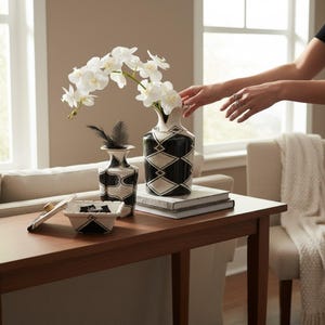 May include: A decorative arrangement featuring two vases and a small dish with a lid. The vases have black and white geometric patterns and hold white flowers and black feathers. The dish is white with black accents.