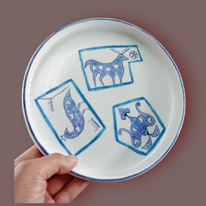 May include: White ceramic plate with blue hand-drawn illustrations. The plate features three distinct designs: a deer, a bird, and an abstract creature, each outlined in blue. The plate has a blue rim.