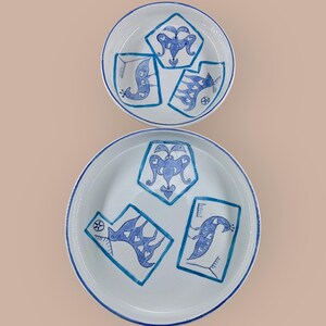 May include: Two white ceramic plates with a blue hand-painted design. The design features stylized birds and geometric shapes. The plates have a blue rim and are set against a neutral background. The plates are of different sizes.