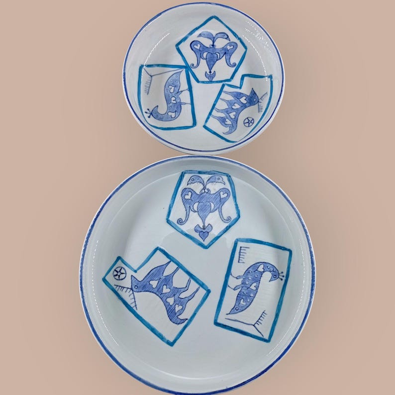 May include: Two white ceramic plates with a blue hand-painted design. The design features stylized birds and geometric shapes. The plates have a blue rim and are set against a neutral background. The plates are of different sizes.