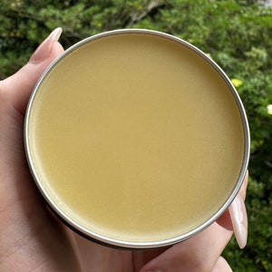 May include: A round, silver-rimmed container filled with a smooth, light yellow balm. The balm has a slightly glossy appearance. The container is held in a hand, with a green, leafy background.