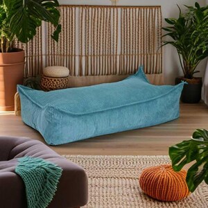 May include: A teal blue corduroy floor cushion with a rectangular shape and a slightly rounded edge. The cushion is sitting on a wooden floor with a rug in front of it.