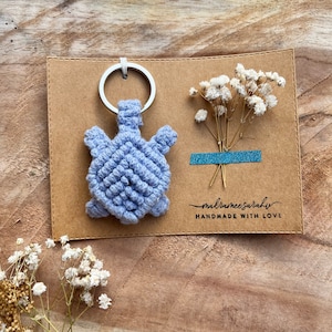 Könnte beinhalten: Ein hellblauer Makramee-Schildkröten-Schlüsselanhänger mit einem silbernen Schlüsselring. Der Schlüsselanhänger befindet sich auf einer braunen Karte mit dem Text "makrameesarah handmade with love".