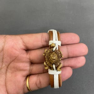 Puede incluir: Una pulsera de cuero marrón con un dije de tortuga. La pulsera tiene una banda de cuero marrón con detalles de cordón blanco. El dije de tortuga es de color marrón claro y está centrado en la pulsera.