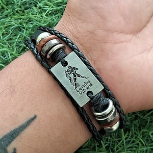 May include: Black braided leather bracelet with a rectangular silver-tone plate featuring the Aquarius zodiac symbol and the dates 1/20-2/18. The bracelet includes silver and brown accents.
