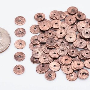 Puede incluir: Un mont&oacute;n de cuentas de metal de color cobre con una superficie ligeramente texturizada. Las cuentas son redondas con un agujero en el centro. Se muestra una moneda de euro para comparar el tama&ntilde;o.