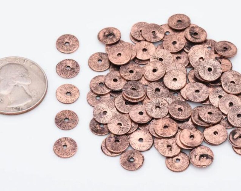 Puede incluir: Un mont&oacute;n de cuentas de metal de color cobre con una superficie ligeramente texturizada. Las cuentas son redondas con un agujero en el centro. Se muestra una moneda de euro para comparar el tama&ntilde;o.