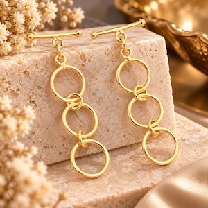 May include: A pair of gold-toned drop earrings featuring a horizontal bar top connected to a vertical chain of three interlocking circular rings. The earrings have a polished metallic finish and a simple, geometric design with a toggle-style closure.
