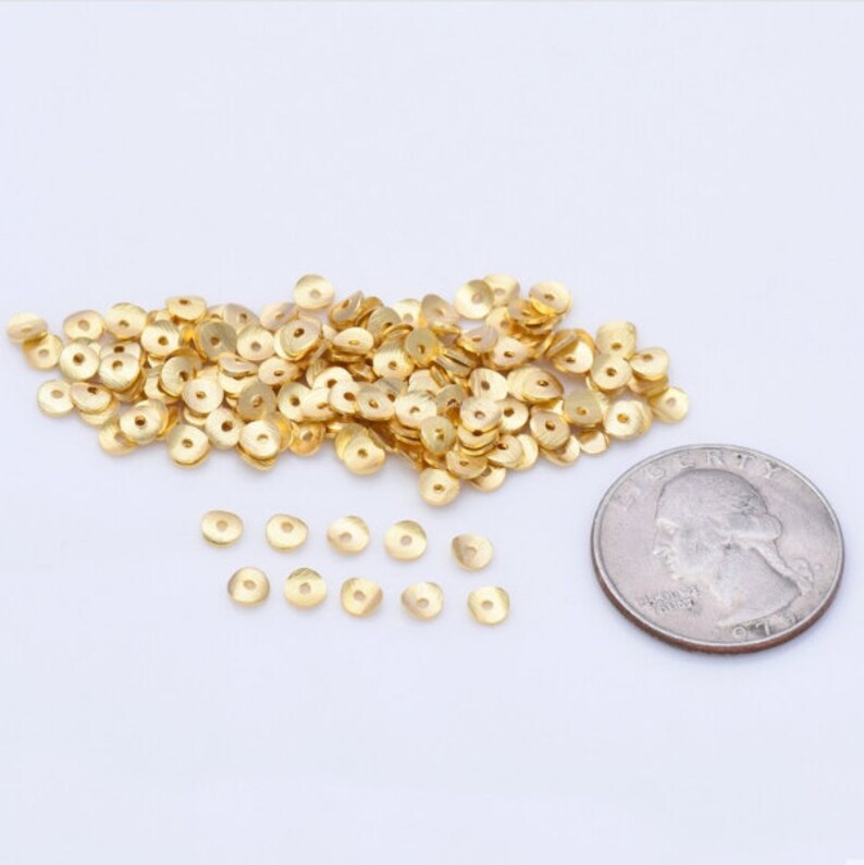 May include: A collection of small, gold-colored metal disc beads with a hole in the center, scattered on a white surface. A U.S. quarter coin is included for size comparison. These beads are likely used for jewelry making or crafting.