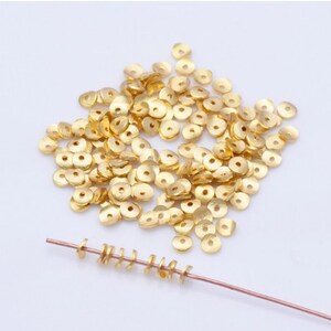 May include: A collection of small, gold-colored, flat, round beads with a central hole, scattered on a white surface. A few beads are strung on a thin, copper-colored wire, showcasing their use in jewelry making or crafts.