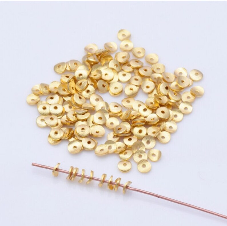 May include: A collection of small, gold-colored, flat, round beads with a central hole, scattered on a white surface. A few beads are strung on a thin, copper-colored wire, showcasing their use in jewelry making or crafts.