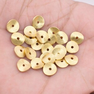 May include: A pile of gold-colored metal beads with a slightly domed shape and a small hole in the center. The beads are arranged in a random pattern on a hand.