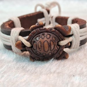 Puede incluir: Pulsera de cuero marrón con un dije de tortuga. La pulsera presenta una banda de cuero marrón oscuro envuelta con cordón blanco. El dije de tortuga es de color marrón claro y está centrado en la pulsera.