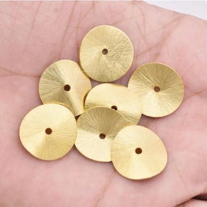 May include: A collection of gold-colored, circular metal components with a textured, radial pattern. Each piece has a small, centered hole. The components are displayed on a light-colored surface, possibly a hand.
