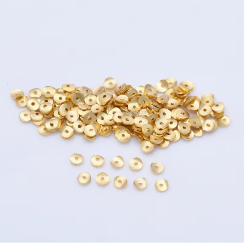 May include: A pile of small, gold-colored metal beads with a flat, disc-like shape and a central hole. The beads are scattered across a white surface, with some beads in focus and others slightly blurred.