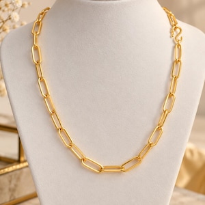 May include: A gold-toned chain necklace featuring elongated, rectangular links with rounded edges. The necklace is secured with a visible S-hook clasp on the right side, showcasing a polished, metallic finish throughout the entire link structure.