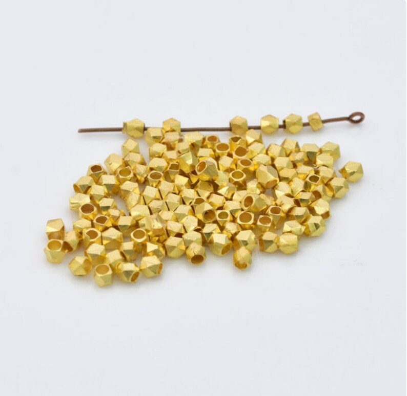 Puede incluir: Un mont&oacute;n de cuentas de metal de color dorado con una forma geom&eacute;trica facetada. Las cuentas est&aacute;n esparcidas sobre una superficie blanca.