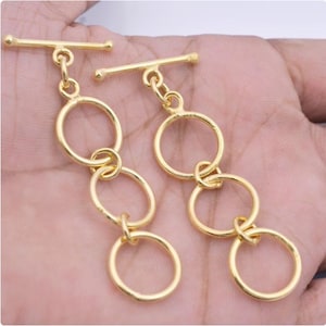 May include: A pair of gold-colored earrings. Each earring features a horizontal bar at the top, connected to three linked circular hoops. The earrings are displayed against a skin-toned background.