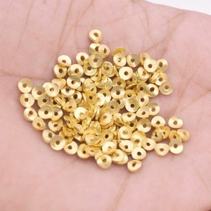 May include: A collection of small, gold-colored, flat, circular beads with a hole in the center. The beads are scattered across a light-colored surface, likely held in a hand. The beads appear to be made of metal and are suitable for jewelry making or crafting.