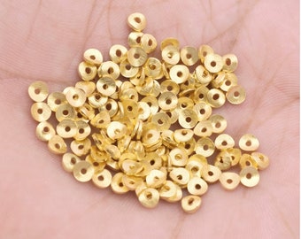 May include: A collection of small, gold-colored, flat, circular beads with a hole in the center. The beads are scattered across a light-colored surface, likely held in a hand. The beads appear to be made of metal and are suitable for jewelry making or crafting.