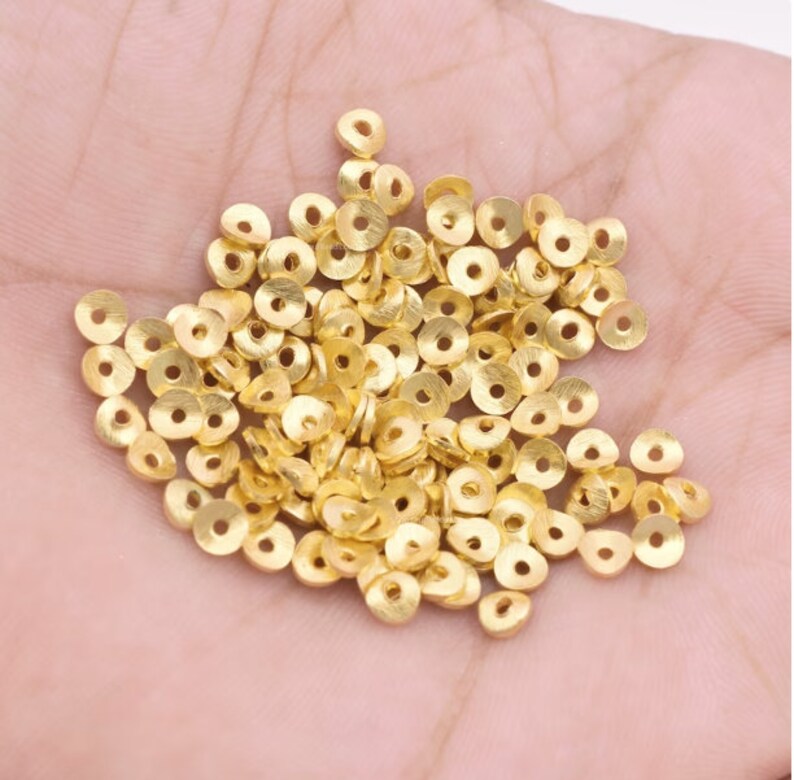 May include: A collection of small, gold-colored, flat, circular beads with a hole in the center. The beads are scattered across a light-colored surface, likely held in a hand. The beads appear to be made of metal and are suitable for jewelry making or crafting.