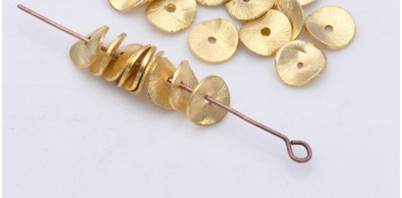 May include: A close-up of a gold wire with a series of gold metal discs strung on it. The discs are flat and have a textured surface. There are several more discs scattered around the wire.