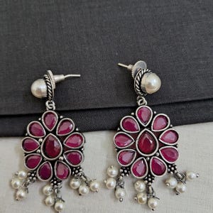 May include: A pair of silver earrings with pink gemstone accents and pearl drops. The earrings feature a floral design with a central pink gemstone surrounded by smaller pink gemstones.