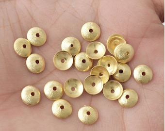 Può includere: Una collezione di piccoli cappucci per perline in metallo color oro. Ogni cappuccio ha un motivo radiale strutturato sulla superficie e un foro centrale. I cappucci per perline sono sparsi su una superficie di colore chiaro.