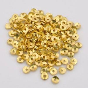 May include: A pile of small, gold-colored metal beads. Each bead is flat, disc-shaped, and has a hole in the center. The beads have a textured surface and a bright, shiny finish, suggesting they are new.