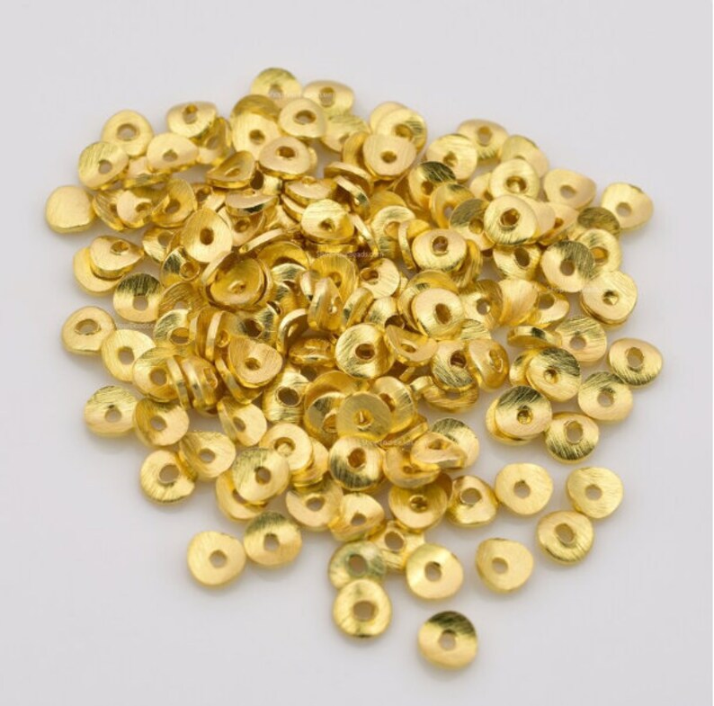 May include: A pile of small, gold-colored metal beads. Each bead is flat, disc-shaped, and has a hole in the center. The beads have a textured surface and a bright, shiny finish, suggesting they are new.