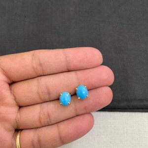 May include: A pair of oval, light blue stud earrings with gold-coloured prongs. The earrings are displayed on a light-coloured surface, with a dark background.