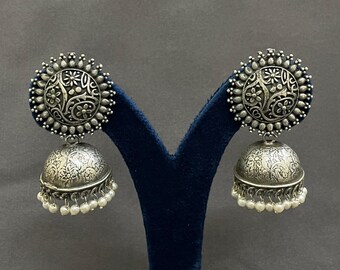 May include: Silver-toned, ornate earrings with a floral design. Each earring features a circular top with a detailed pattern and a bell-shaped bottom adorned with small pearls. The earrings are displayed on a dark blue stand.