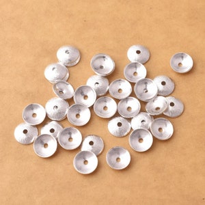 May include: A group of silver-toned metal bead caps, scattered on a brown surface. The caps are round with a raised centre and a hole in the centre.