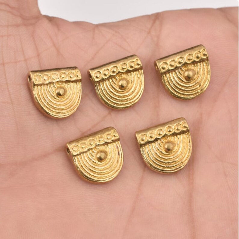 May include: Five gold-toned metal pendant charms with a rounded, half-circle shape. Each charm has a raised, textured design with concentric circles and a small, round bead in the center.