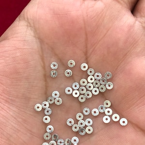 Puede incluir: Un conjunto de pequeñas cuentas circulares, planas y plateadas, con un agujero en el centro. Las cuentas tienen una superficie texturizada y están dispersas sobre un fondo claro. Adecuadas para la creación de joyas u otros proyectos de manualidades.