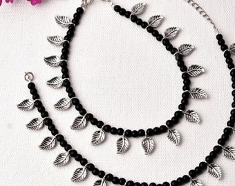 May include: Two anklets with black beads and silver-colored leaf charms. The anklets are displayed on a white surface, with a sprig of pink flowers in the background. The anklets have adjustable chains.