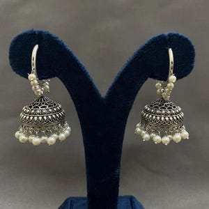 Puede incluir: Pendientes plateados en forma de campana, con intrincados detalles de filigrana y pequeñas perlas blancas colgantes. Los pendientes se exhiben en un soporte de terciopelo azul oscuro, mostrando su diseño ornamentado.