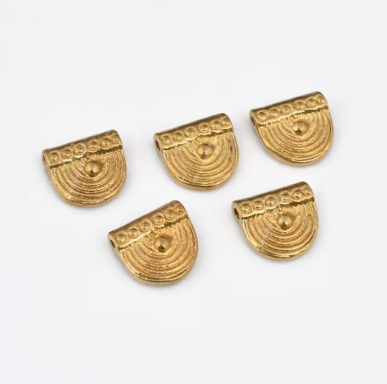May include: Five gold-colored metal charms with a decorative design. Each charm features a half-circle shape with a raised center and a beaded border.