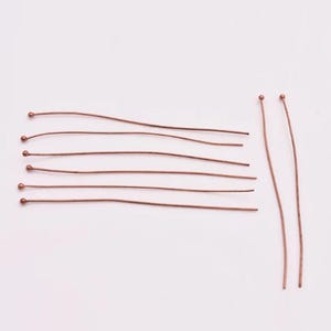 May include: A set of ten copper head pins with rounded heads. The pins are arranged in two rows, with five pins in each row. The pins are all the same length and have a shiny copper finish.